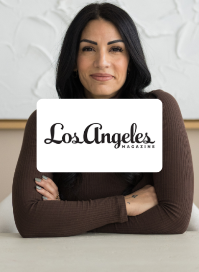 Portrait of a woman seated at a white table with arms crossed, logo overlay reading Los Angeles Magazine in the center (studio backdrop).